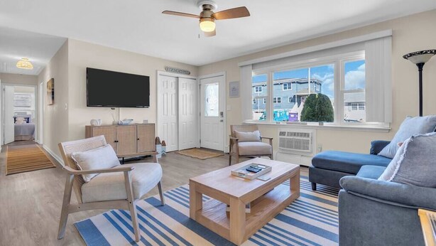A modern living room with a flat-screen TV, a ceiling fan, and a large window with a view of buildings. - Professionally Managed | Walk to Beach (Seaside Heights)