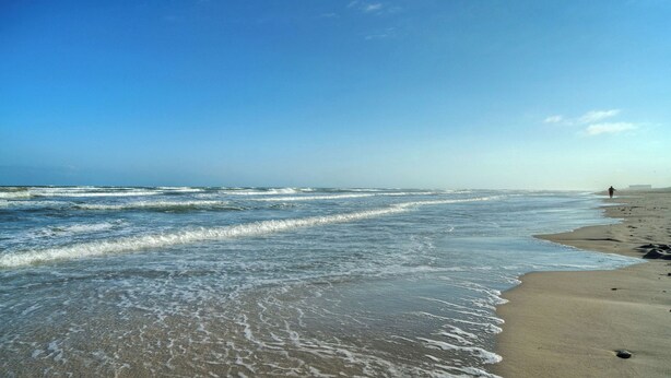 A beach with waves, a person walking, and a clear sky. - Stylish 2BR Coastal Getaway w/ Pool & Prime Location Lazy Fun (Port Aransas)