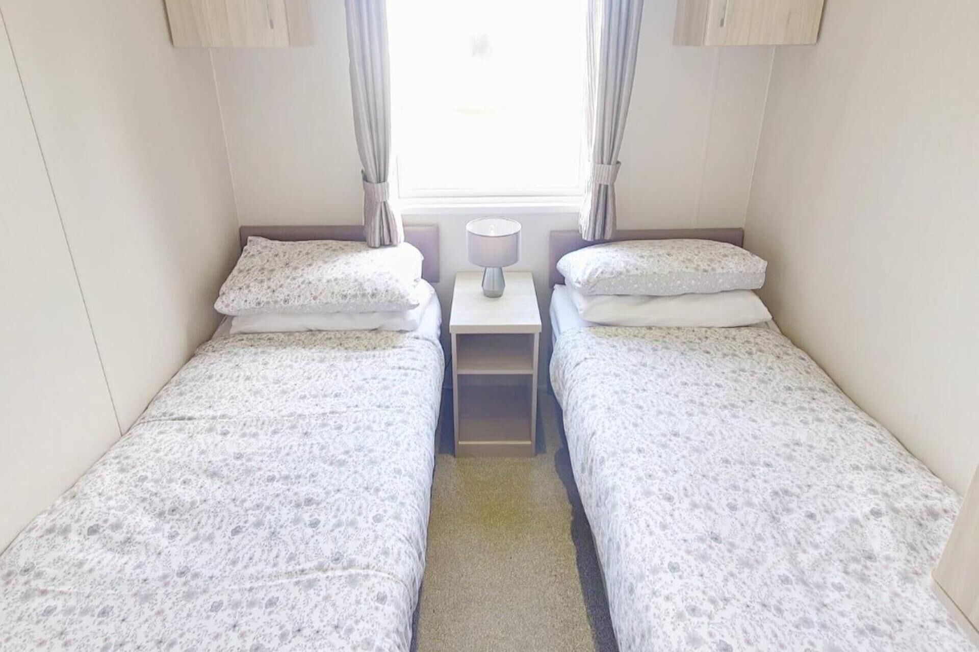 Une petite chambre d’hôtel composée d’une seule pièce, avec deux lits, une table de chevet, une lampe et une fenêtre avec des rideaux.