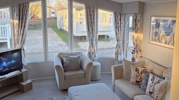 Un salon moderne avec un canapé, un fauteuil et un téléviseur à écran plat. De grandes fenêtres offrent une vue sur une cour où se trouvent des maisons mobiles.