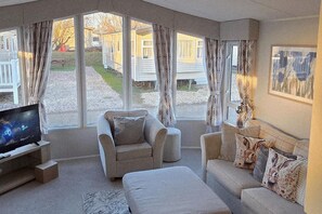 Una sala de estar moderna con un sofá, un sillón y un televisor de pantalla plana. Amplias ventanas ofrecen vistas a un patio con casas móviles.