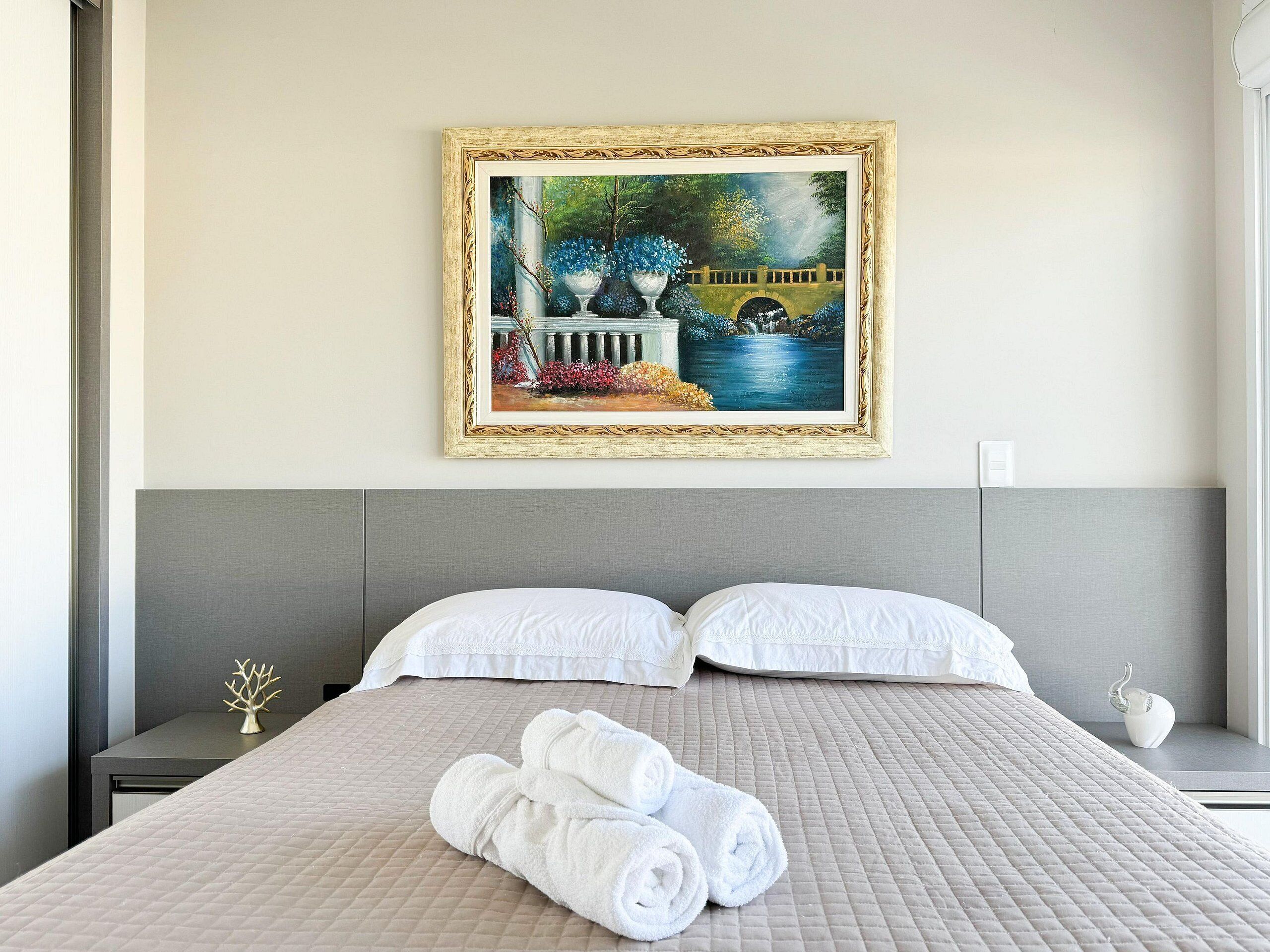 A neatly made bed with white linens, a painting of a garden scene on the wall, and a small decorative tree on the nightstand.