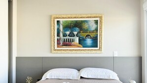 Una cama bien hecha con sábanas blancas, un cuadro de una escena de jardín en la pared y un pequeño árbol decorativo en la mesita de noche.