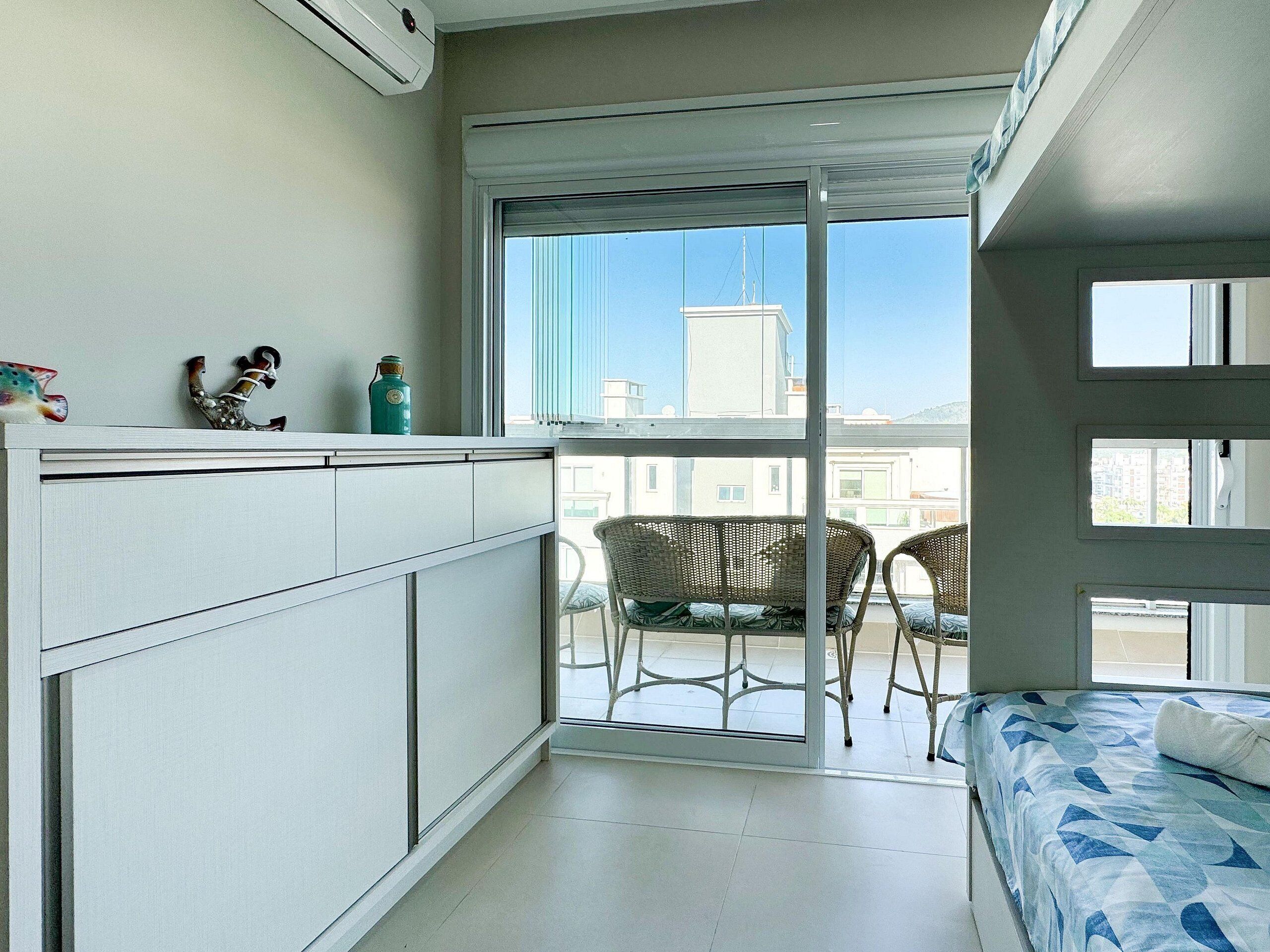 Quarto compacto com cama, pequeno espaço de cozinha, varanda com cadeiras e vista para prédios.