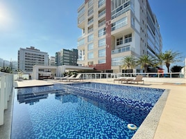 Uma piscina com borda azulejada, rodeada por espreguiçadeiras e um edifício com várias varandas.