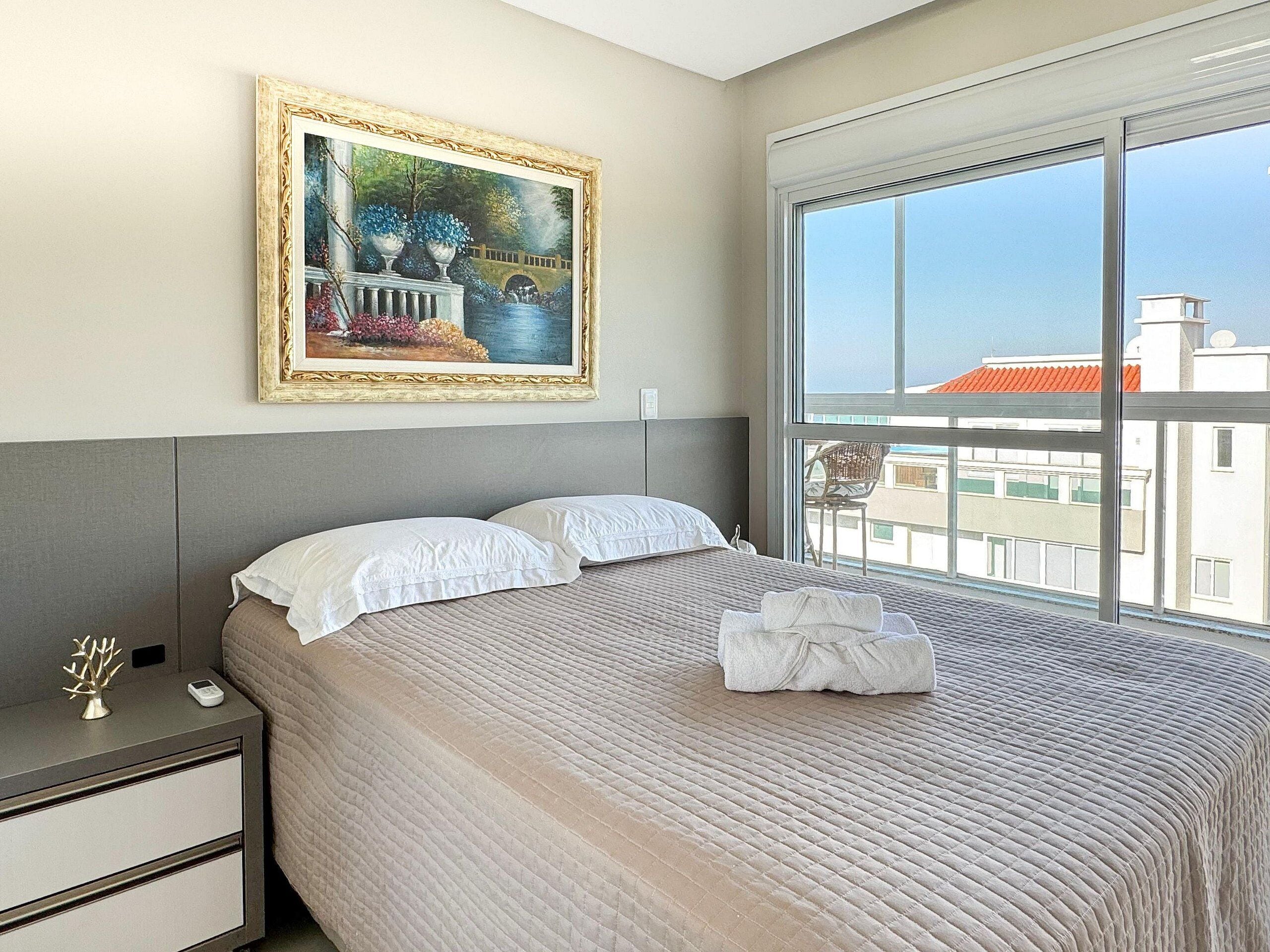 A hotel room with a large window, a bed with white bedding, a nightstand, and a painting on the wall.
