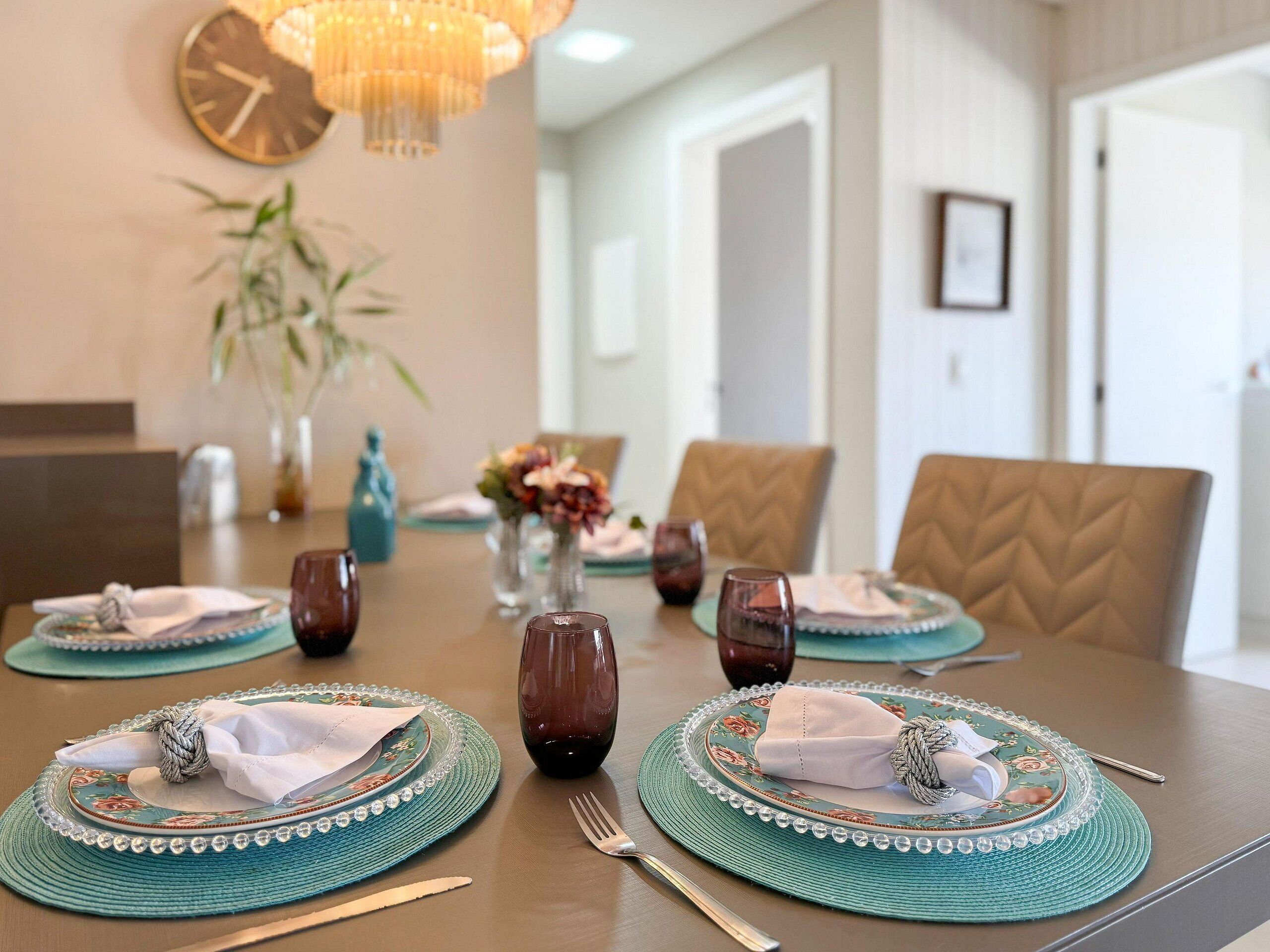 A dining table set with plates, cutlery, and napkins, with a clock on the wall and a chandelier above.