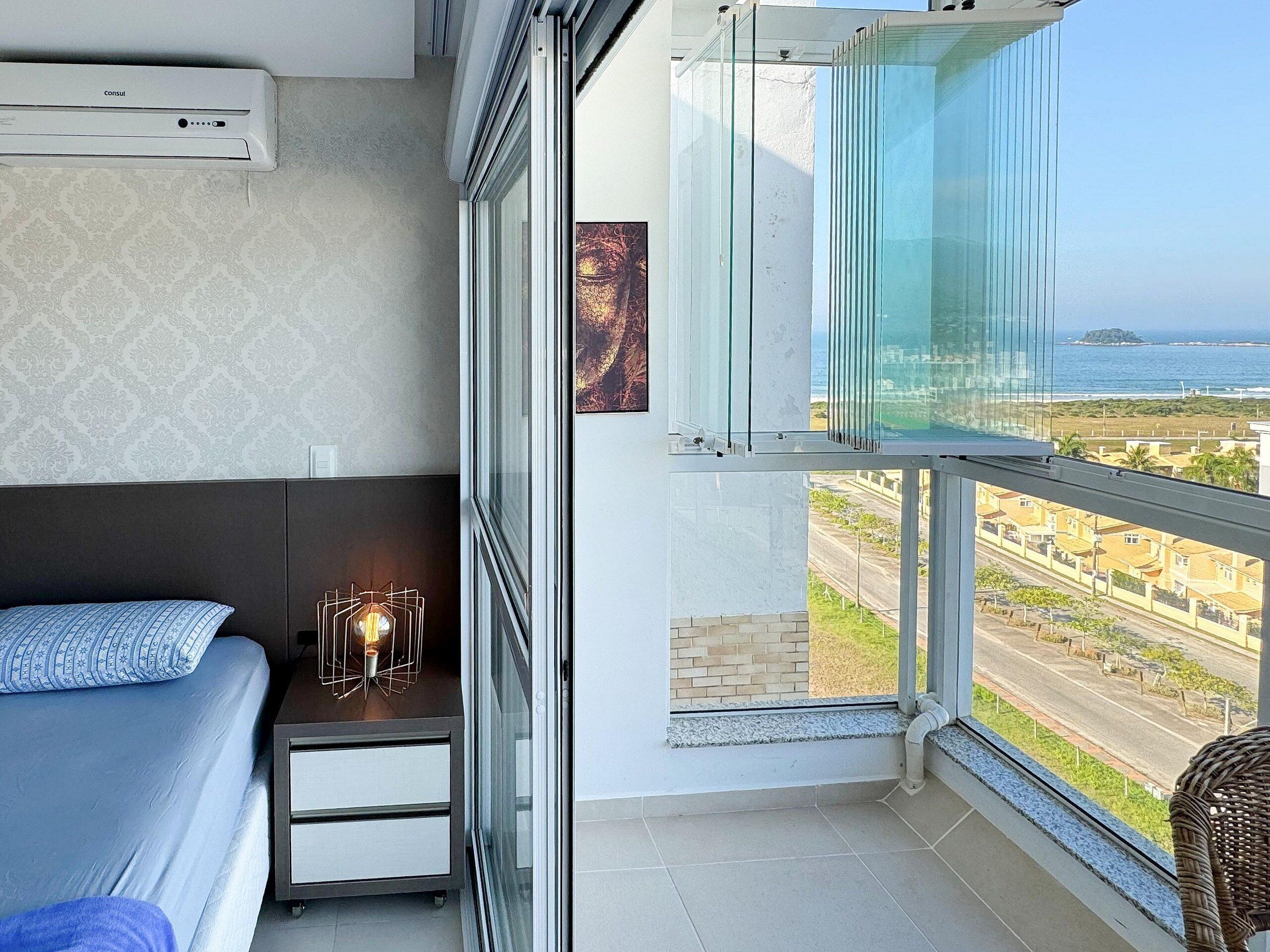 Un balcón con vista al mar y edificios, una cama con un cojín estampado, una mesita de noche con una lámpara y una ventana con barandilla de cristal.