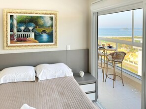 A bedroom with a bed, a nightstand, a painting, and a balcony with a view of the sea.