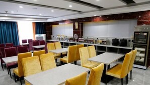 A modern dining area with yellow chairs and white tables, a buffet counter, and a kitchen area with ovens.