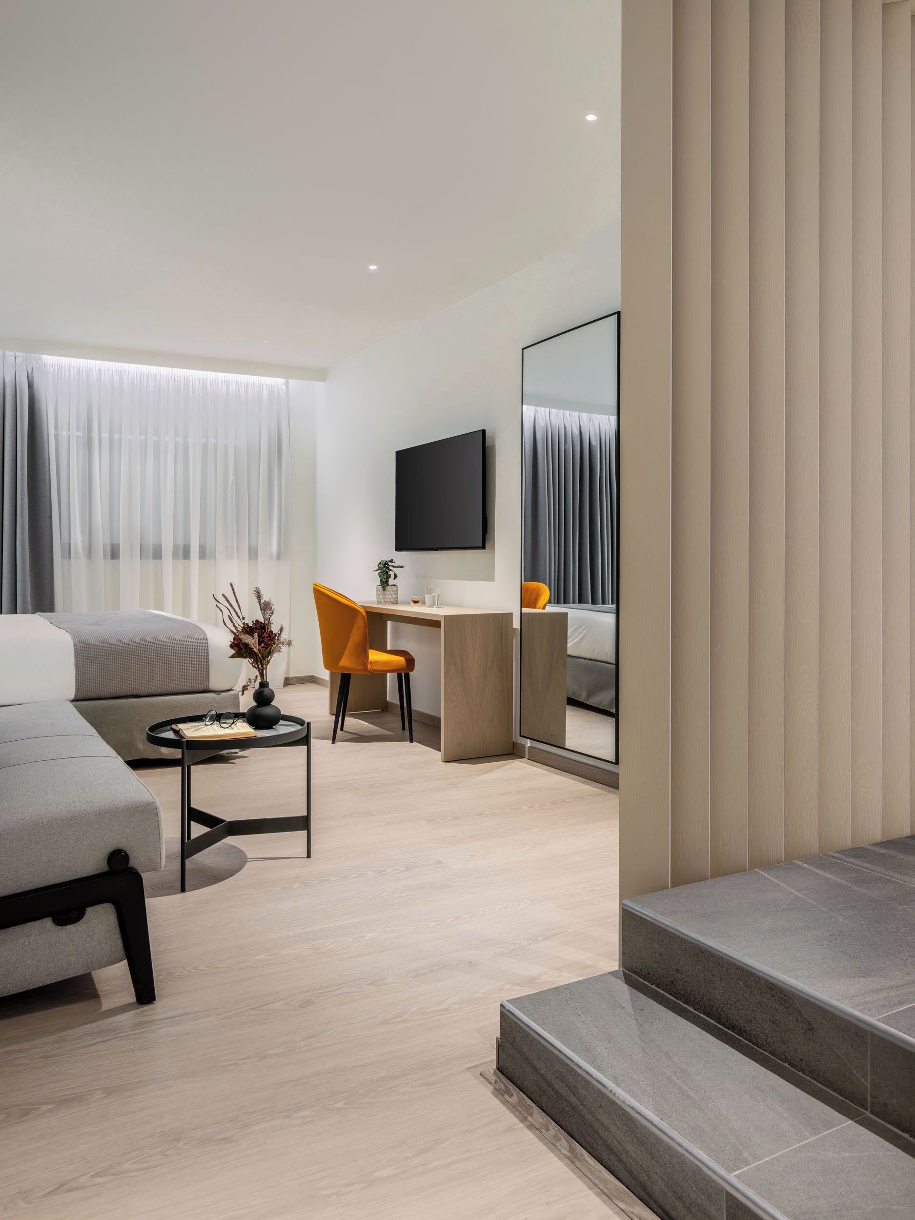 A modern hotel room with a sofa, a desk, a chair, a TV, and a large mirror.