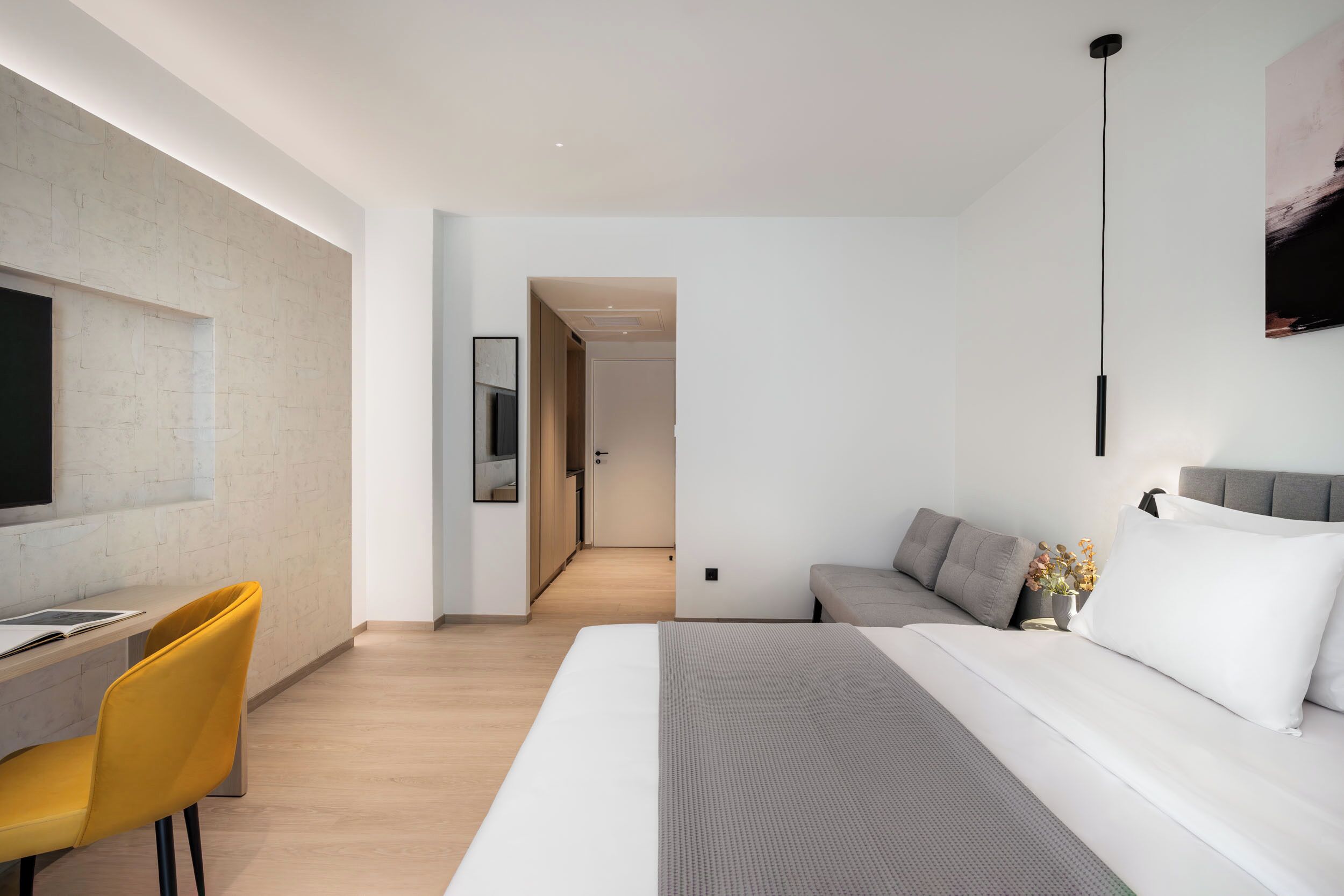 A modern hotel room with a bed, a desk, and a chair. There is a hallway leading to another room.