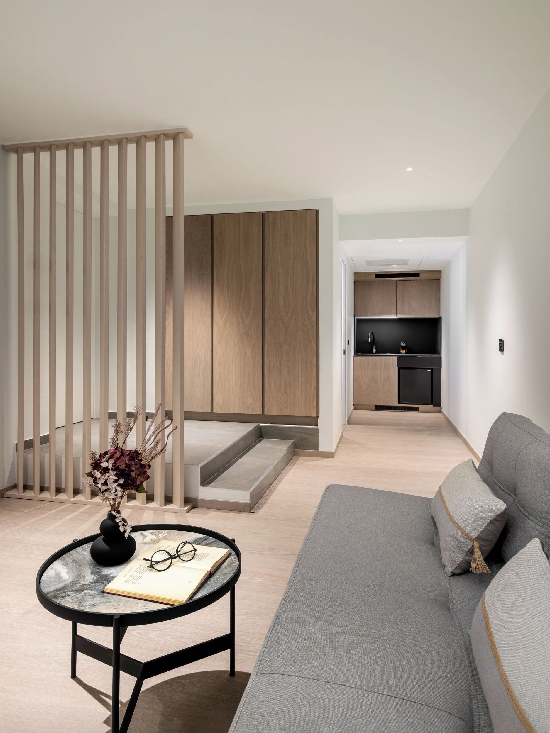 Eine moderne Wohnung mit einem grauen Sofa, einem kleinen runden Tisch mit einer Vase und Gläsern, einer Treppe mit Holzvertäfelung und einer Küchenzeile im Hintergrund.