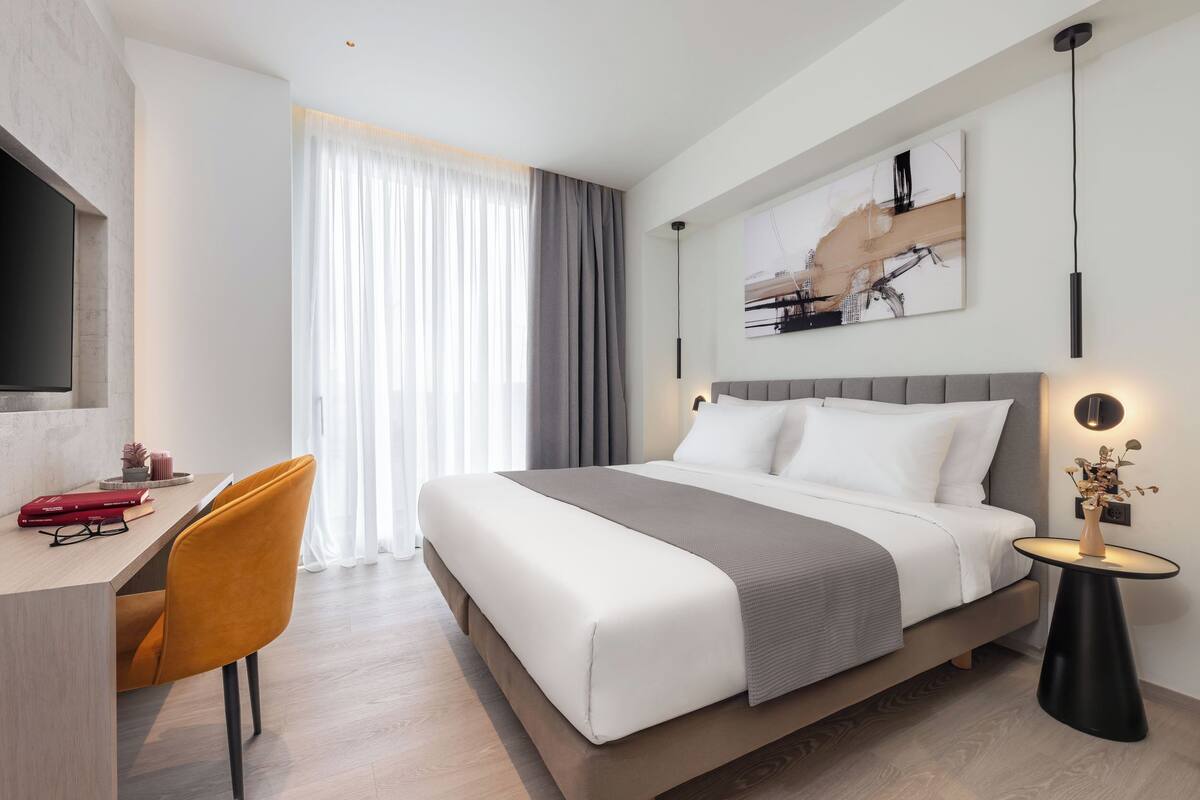 A modern hotel room with a large bed, a desk with a chair, a flat-screen TV, and a painting on the wall.