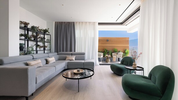 A modern living room with a grey sectional sofa, a green armchair, and a coffee table. There is a black shelving unit with plants, a view of the outdoors through large windows, and a wooden accent wall. - Urban-K (Athens)