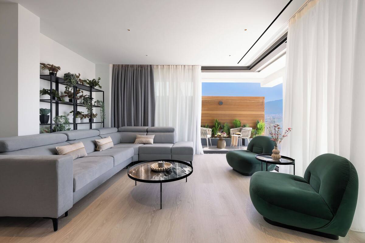 A modern living room with a grey sectional sofa, a green armchair, and a coffee table. There is a black shelving unit with plants, a view of the outdoors through large windows, and a wooden accent wall.