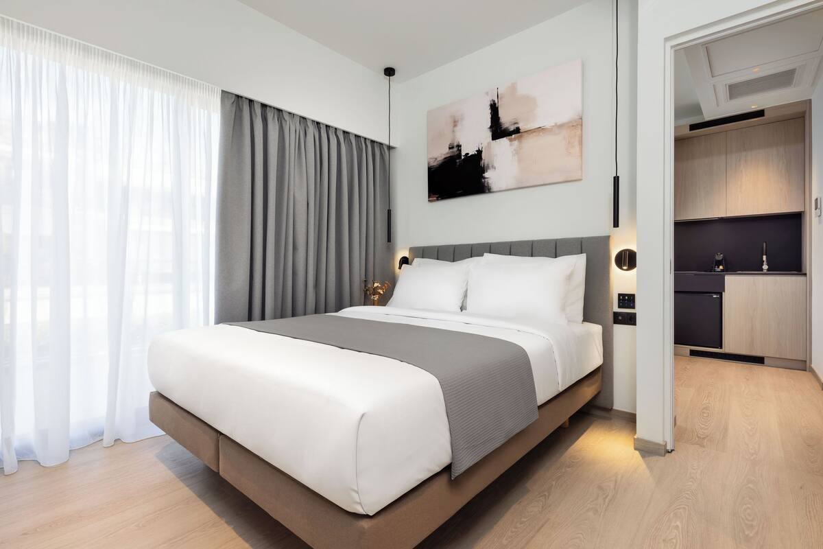 A modern hotel room with a large bed, a view of the cityscape, a kitchen area, and a dining table.