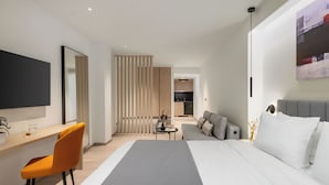 A modern hotel room with a large bed, a desk with a chair, a sofa, a TV, and a kitchen area in the background. - Urban-K (Athens)