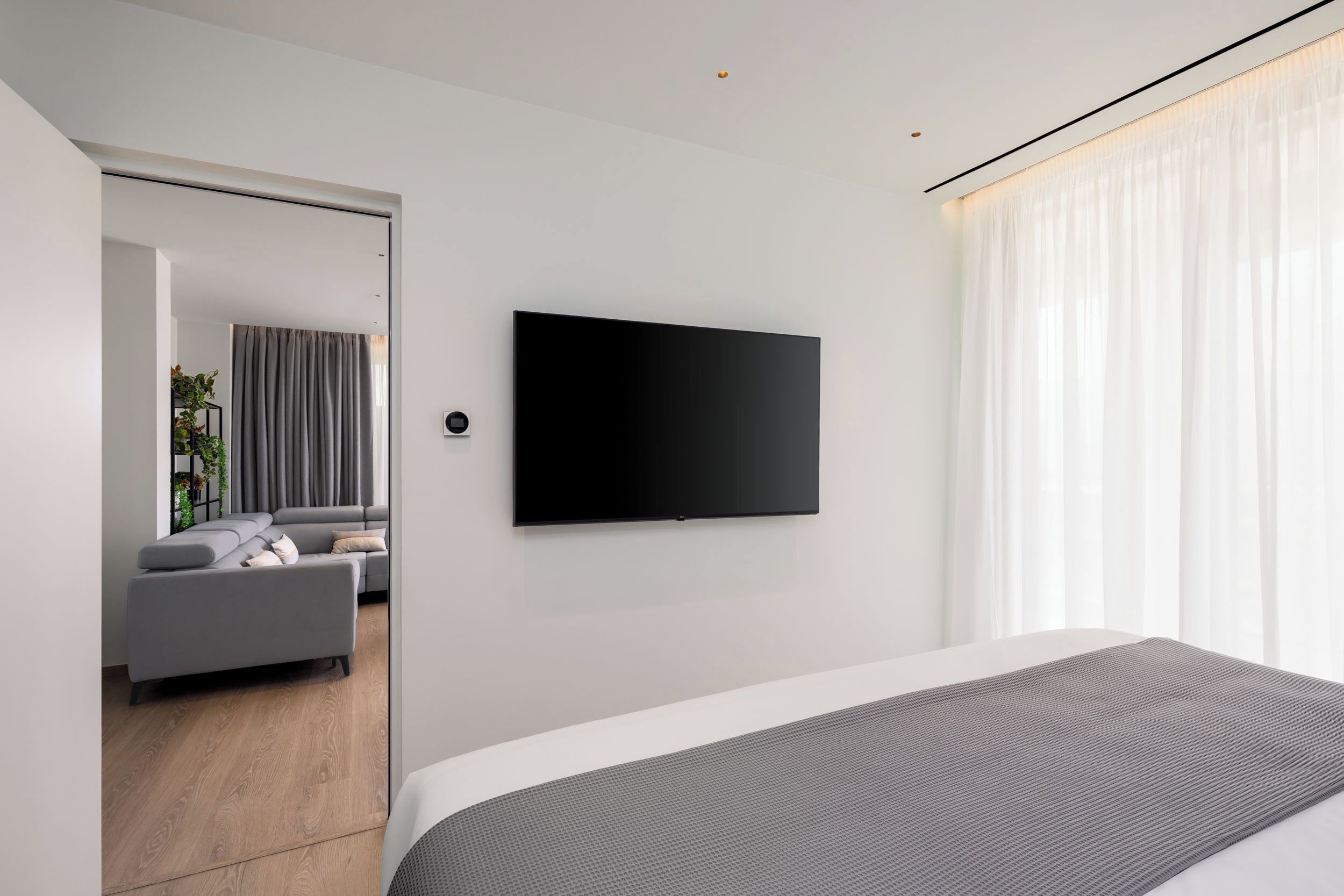 A modern bedroom with a large bed, a flat-screen TV mounted on the wall, and a view of a living area with a sofa and curtains.