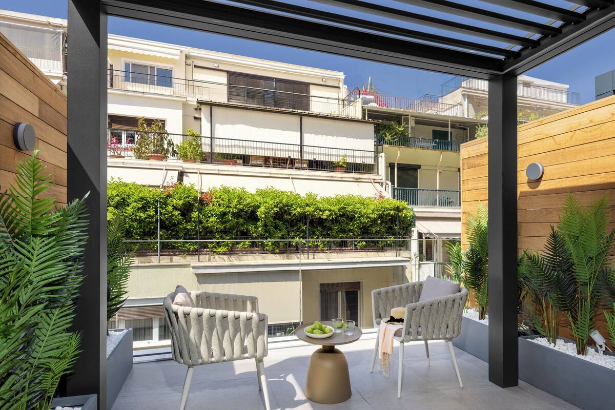 A covered outdoor patio with two chairs and a table, surrounded by white pebbles and green plants.