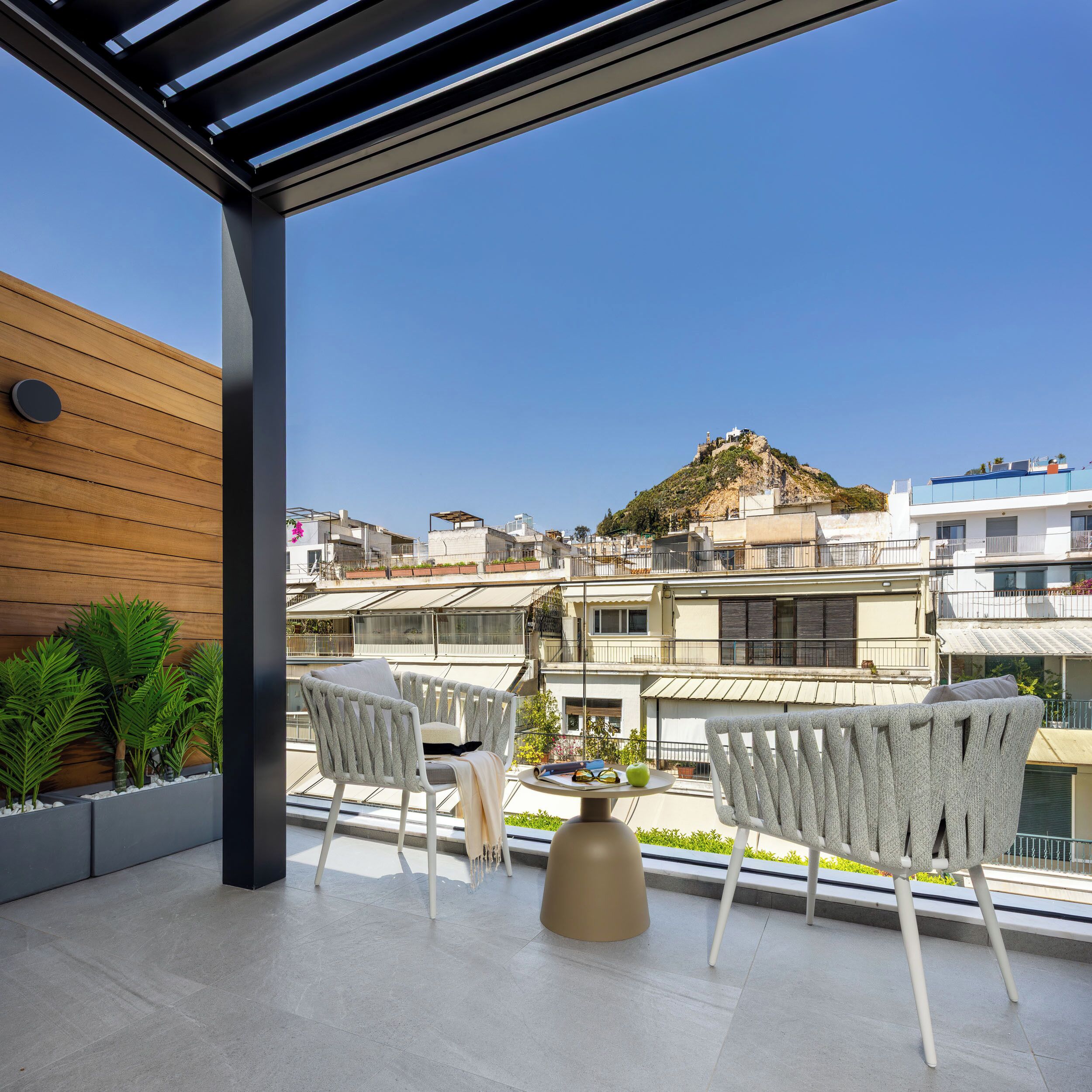 A rooftop terrace with two chairs and a table, overlooking a cityscape with buildings and a hill.