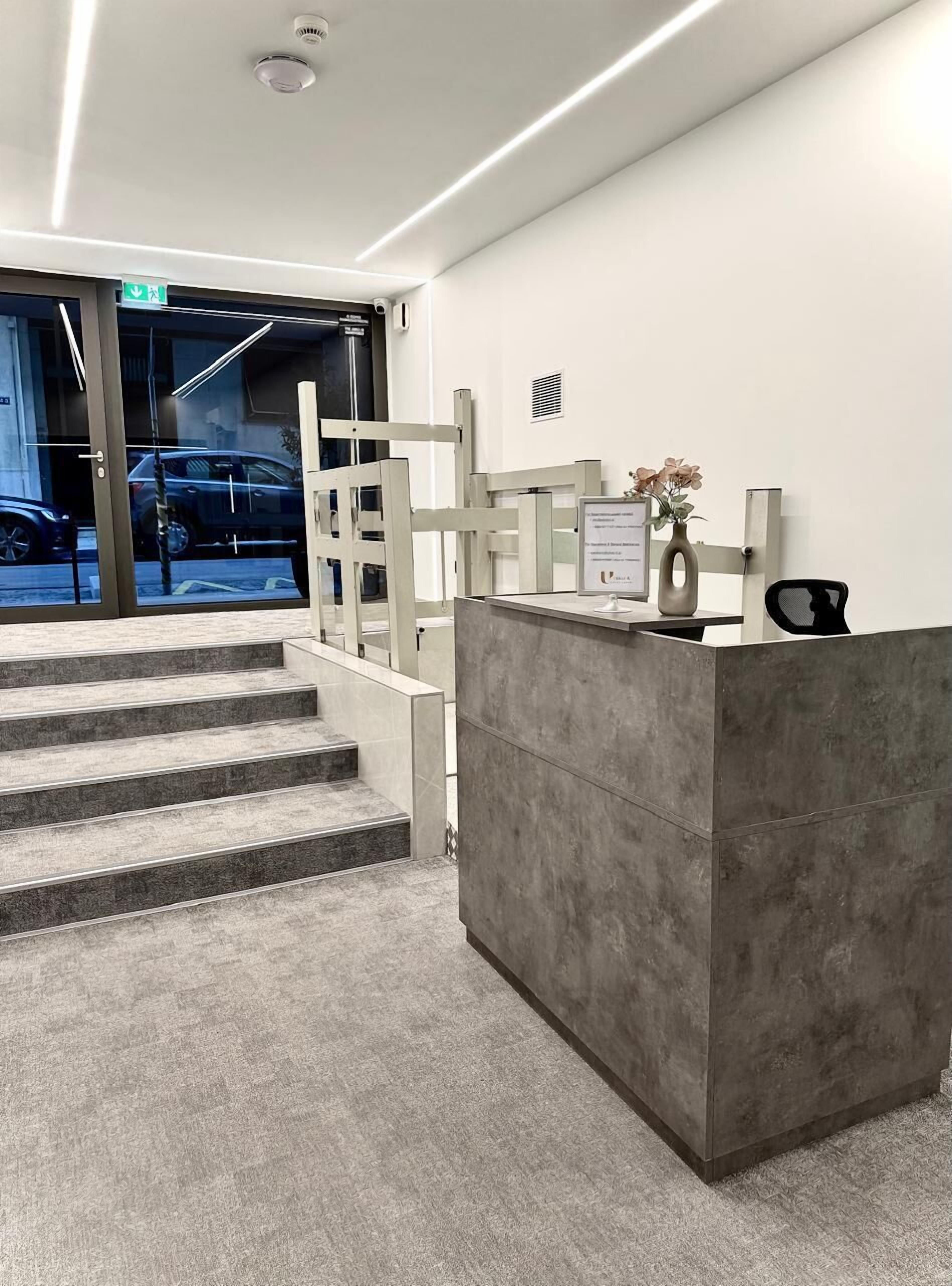 A reception desk with a grey counter, a small vase with flowers, and a staircase leading to an entrance with glass doors.