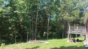 A backyard with a stone fire pit, a wooden deck, and a dense forest in the background. - Charming 1-bedroom cottage with WiFi and AC in lovely Stamping ky (Stamping Ground)