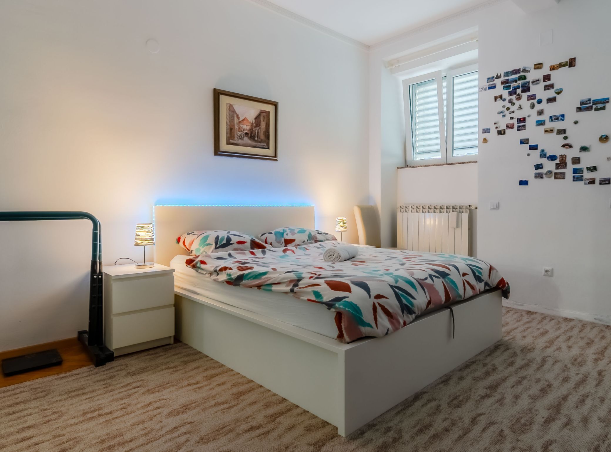 A bedroom with a bed, two bedside tables, a nightstand, a radiator, a window with blinds, and a wall with photos.