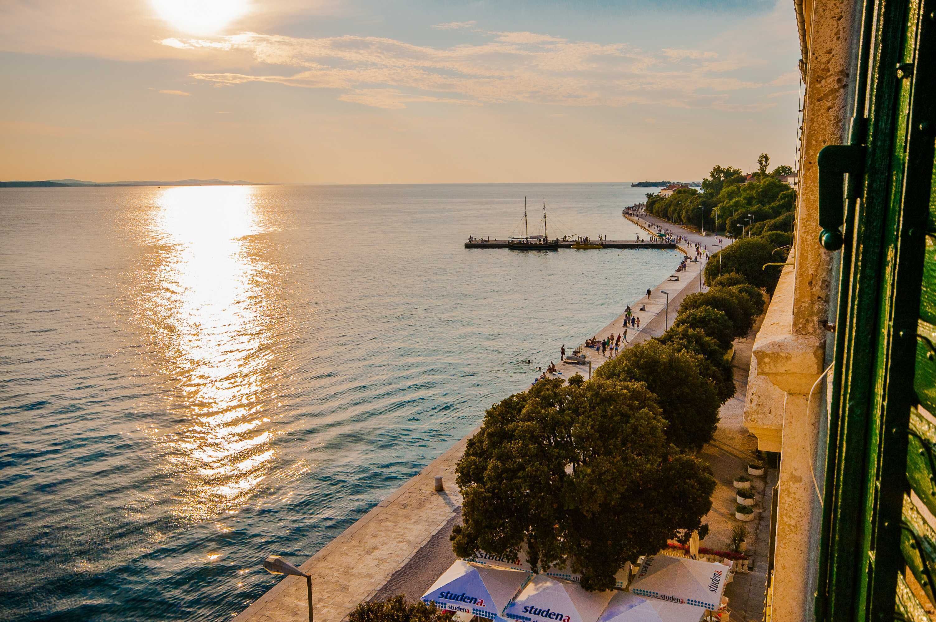 Vista costera con un muelle que se adentra en el mar, un paseo marítimo bordeado de árboles y un edificio de fachada verde.