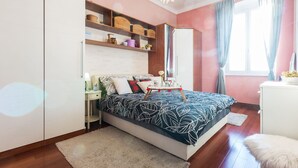 Une chambre à coucher comprenant un lit, une table de chevet, une armoire, des étagères avec des paniers, un miroir et une fenêtre avec des rideaux.