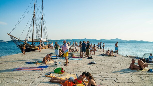 Une plage où des gens se prélassent au soleil et se détendent, un voilier, et une vue dégagée sur la mer et les montagnes lointaines.