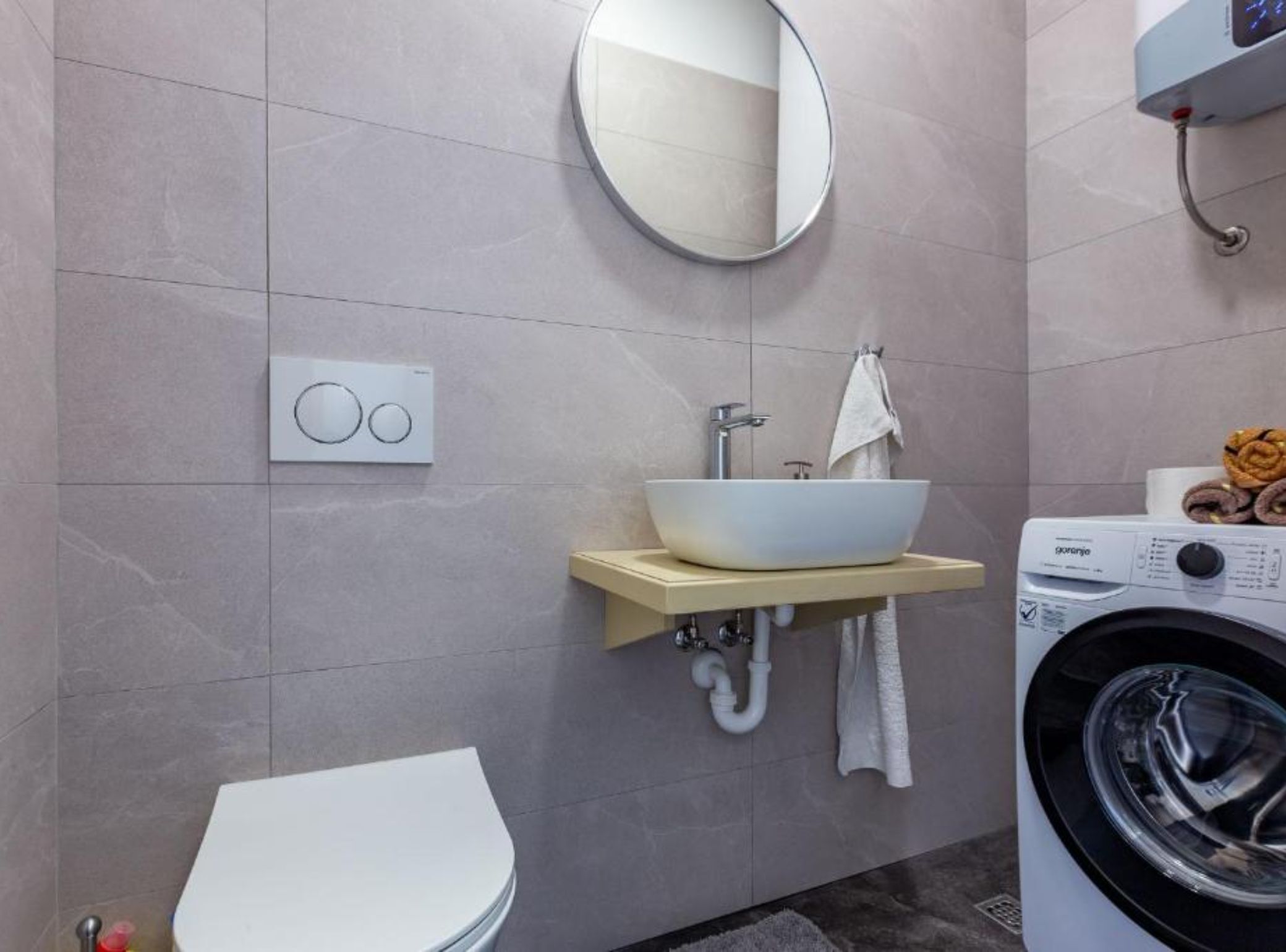 Un baño moderno con lavamanos, espejo y retrete blancos. Hay una lavadora y un perchero con toallas.