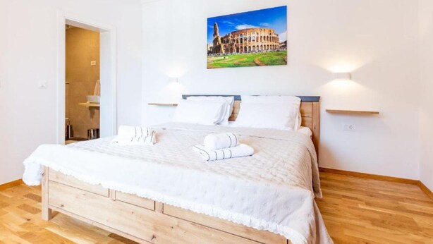 Un dormitorio con cama de madera, ropa de cama blanca, un cuadro del Coliseo y dos luces fijadas a la pared.