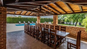 Un patio al aire libre con piscina, mesas y sillas de madera, y una pared de ladrillo.