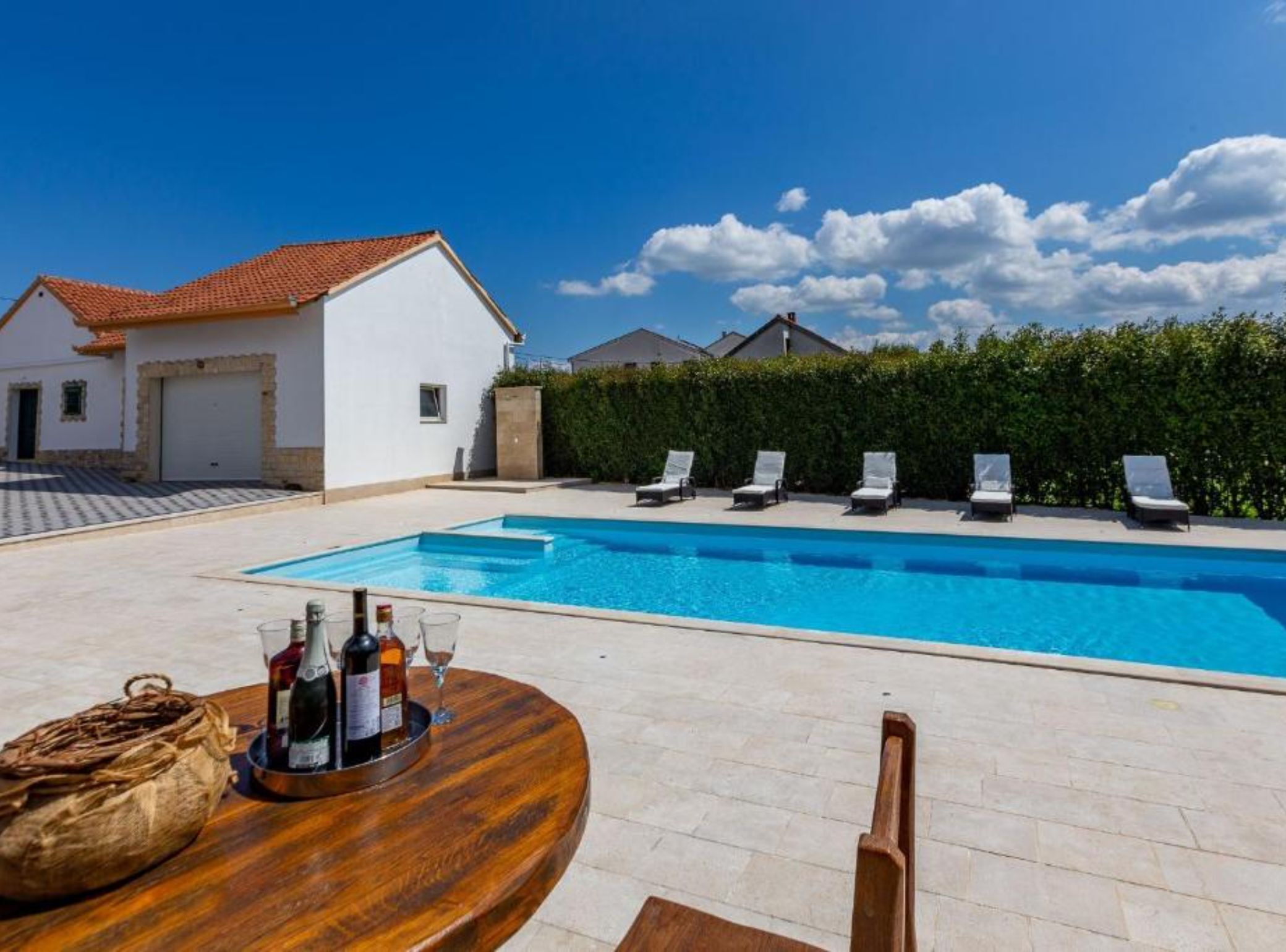 A swimming pool with lounge chairs, a table with bottles and glasses, and a white building with a red roof.