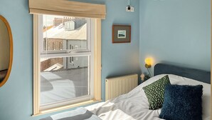 A bedroom with a large window, a radiator, a bedside table with a lamp, and a bed with pillows.
