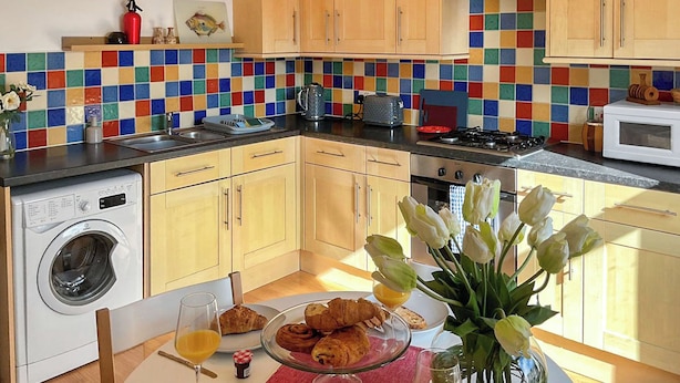 A kitchen with wooden cabinets, a colorful backsplash, a washing machine, and a breakfast table with croissants and tulips.