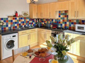 Une cuisine avec des meubles en bois, un crédence aux couleurs vives, un lave-linge et une table de petit-déjeuner avec des croissants et des tulipes.