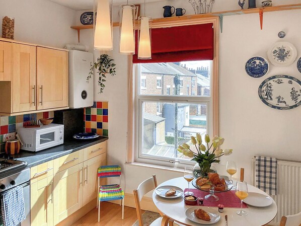 Une cuisine avec des meubles en bois, un four à micro-ondes, un plan de travail coloré recouvert de carreaux, une fenêtre avec un store rouge et un coin repas avec une table mise pour le petit-déjeuner.