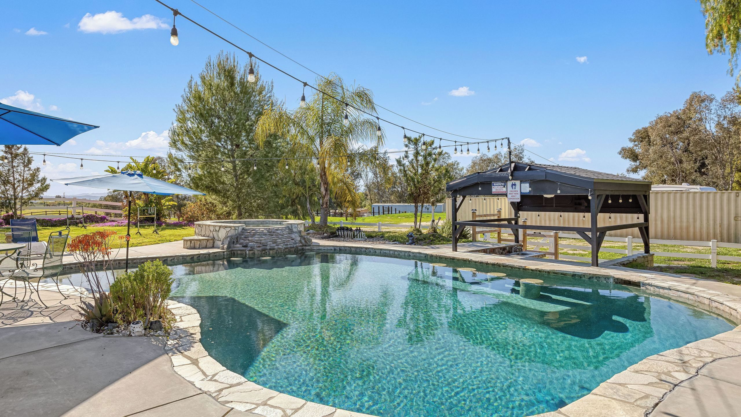A clear swimming pool with a stone border, a wooden deck, and a gazebo.