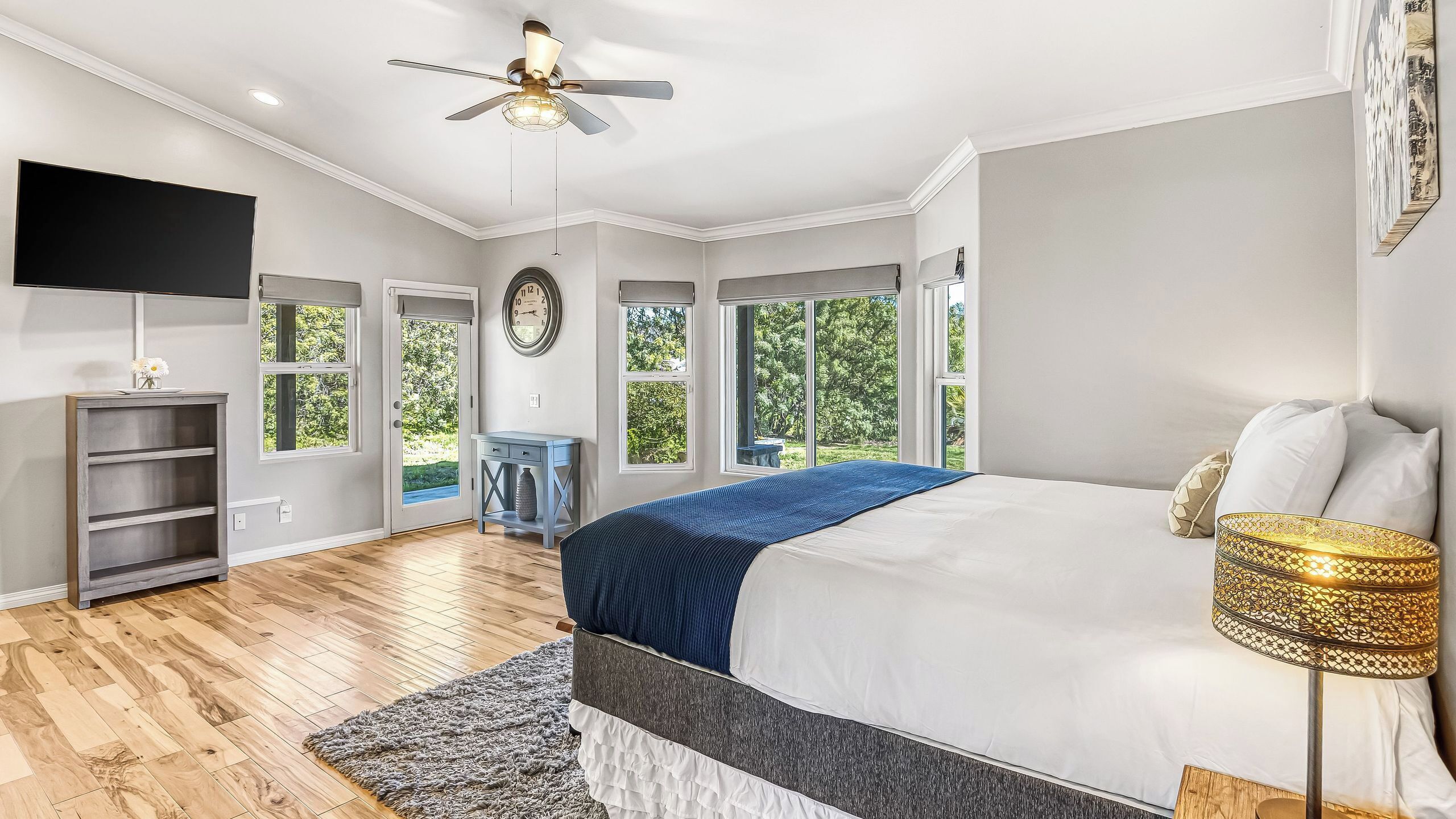 A bedroom with a bed, a TV, a ceiling fan, and large windows.