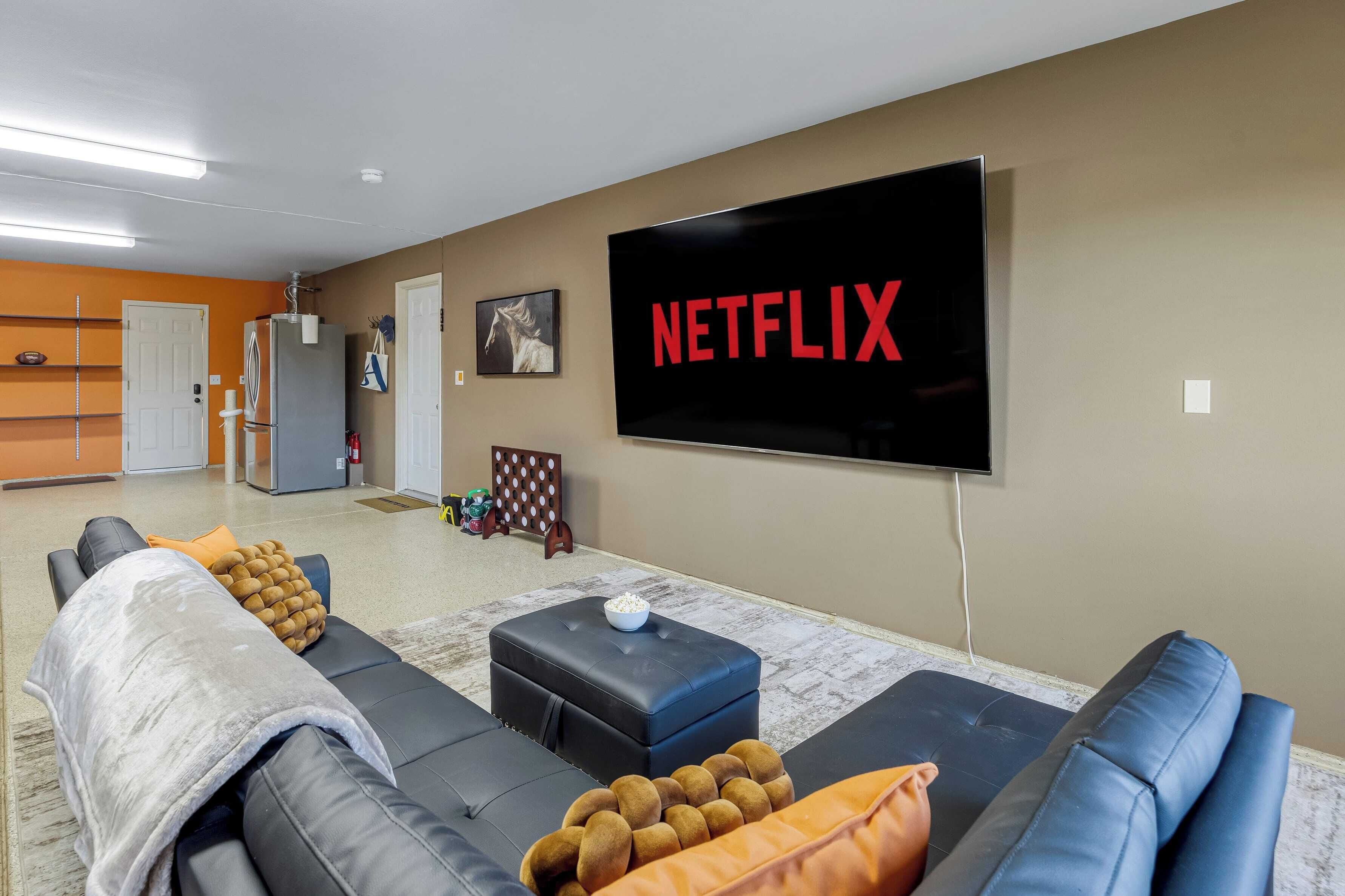 Een woonkamer met een grote televisie waarop 'NETFLIX' staat, een bank, een poef en een klein tafeltje met een schaal.