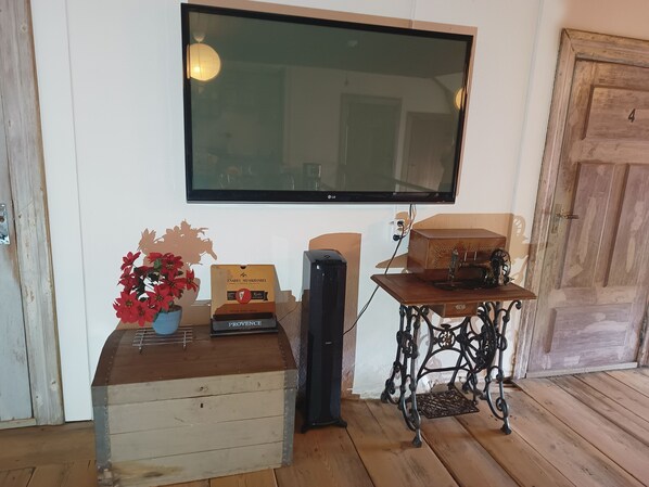 Un cofre de madera con un jarrón de flores rojas, una máquina de coser antigua sobre una mesa y un televisor fijado a la pared.