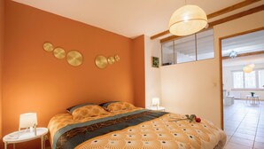 A bedroom with an orange wall, a bed with patterned bedding, a bedside table with a lamp, and a view of a dining area through a glass door.