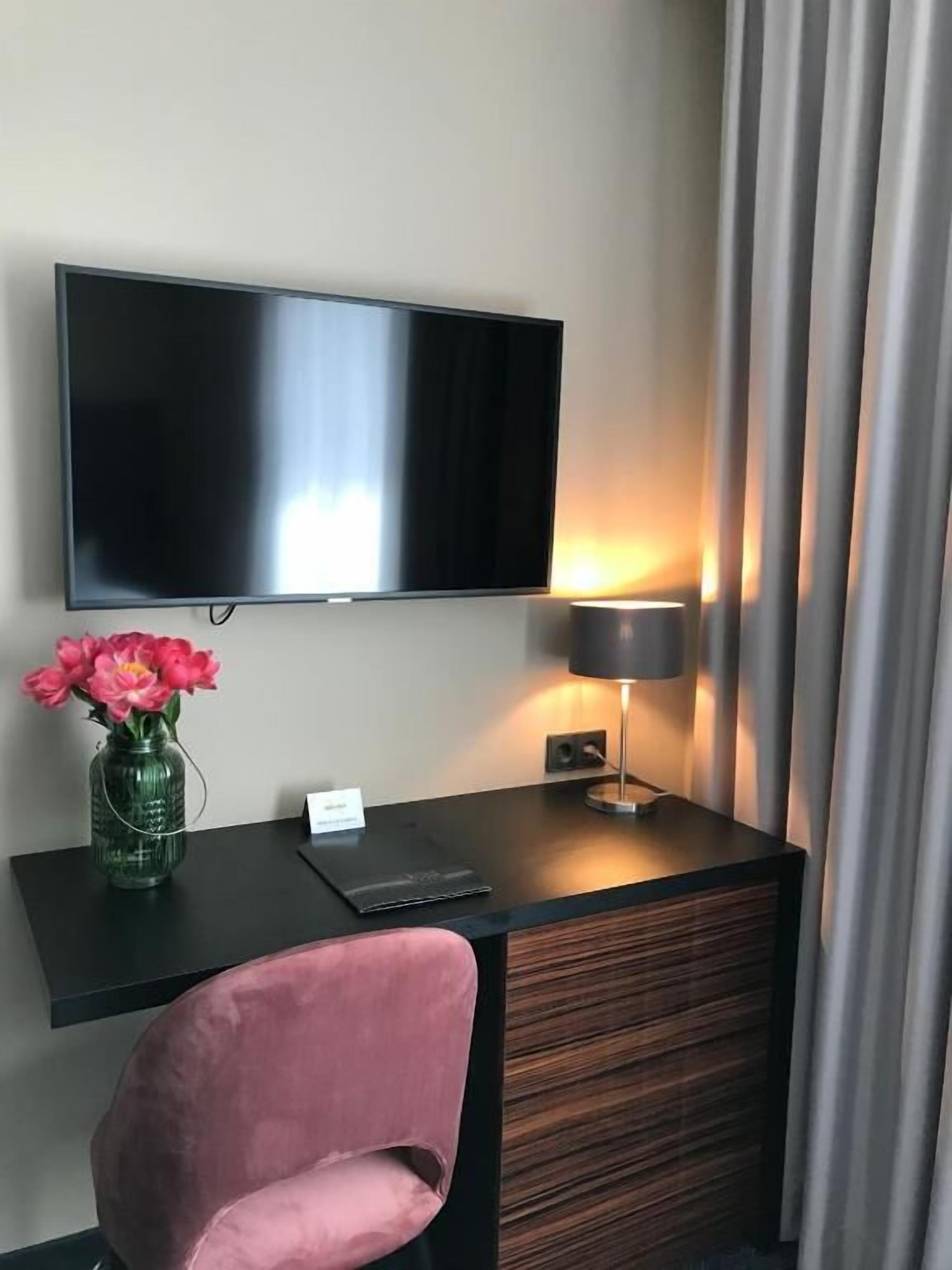 A hotel room with a flat-screen TV mounted on the wall, a desk with a lamp, a vase with flowers, and a pink chair.