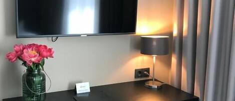 A hotel room with a flat-screen TV mounted on the wall, a desk with a lamp, a vase with flowers, and a pink chair.