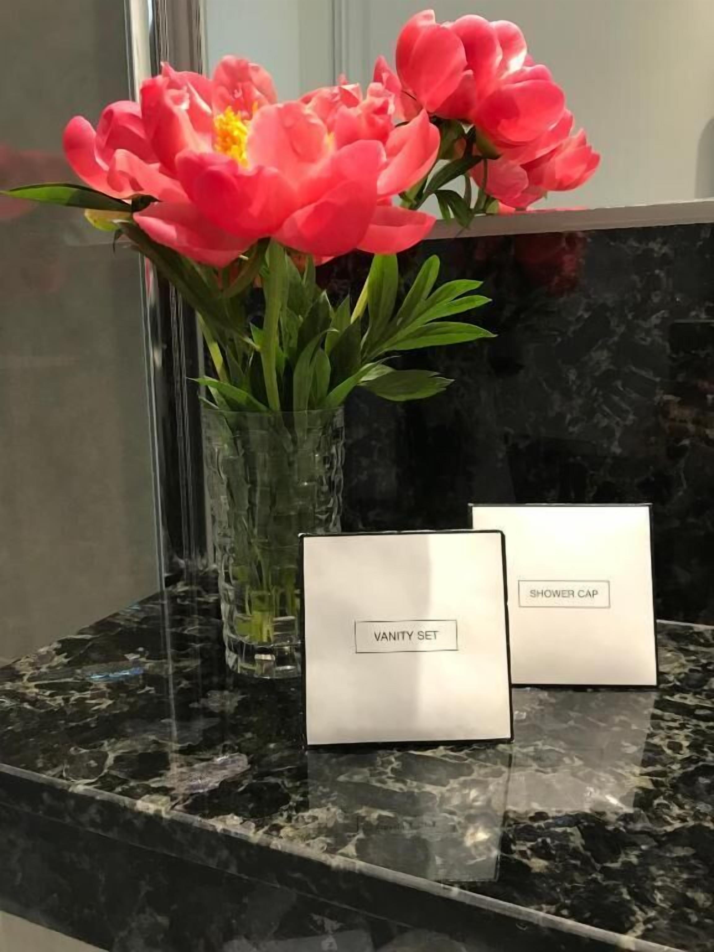 A bathroom vanity with a clear glass vase containing pink flowers, a vanity set, and a shower cap on a marble countertop.