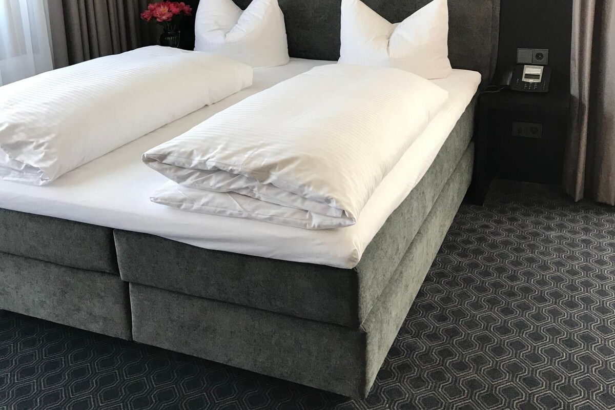 Ein Doppelbett mit grau gepolstertem Kopfteil und Fußteil, weißer Bettwäsche und einem Nachttisch mit Telefon.