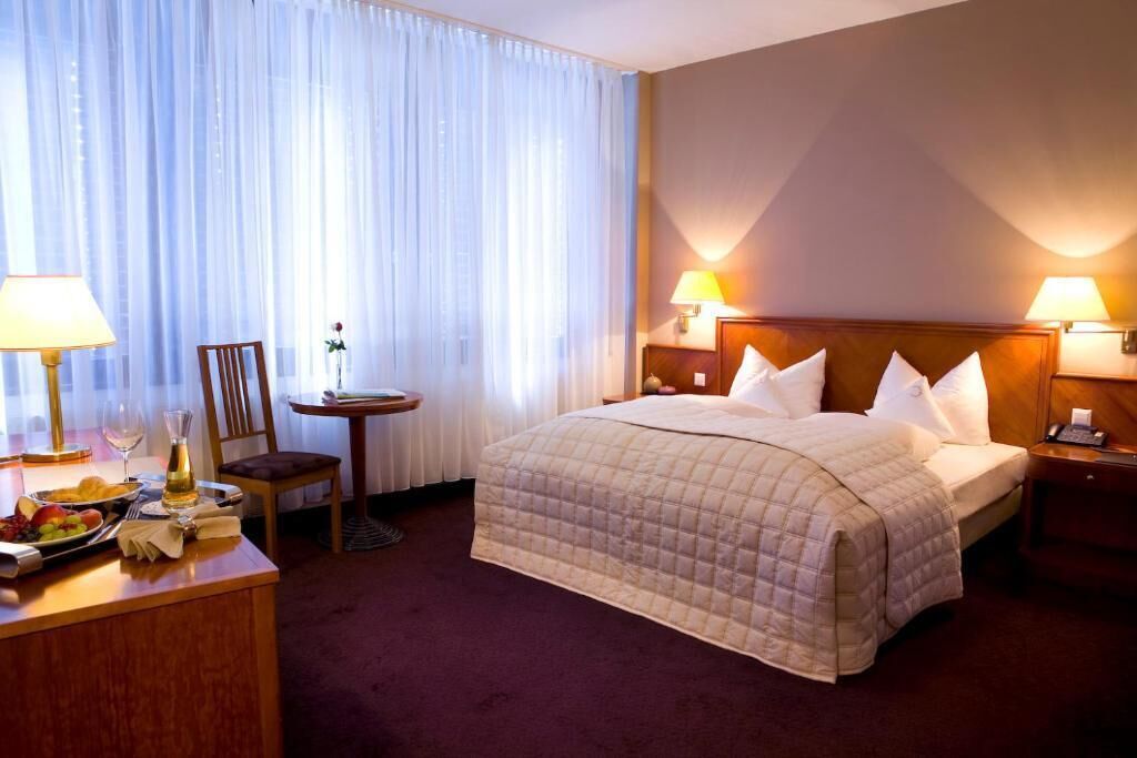 A hotel room with a large bed, two bedside lamps, a small table with a chair, and a desk with a lamp and fruit.