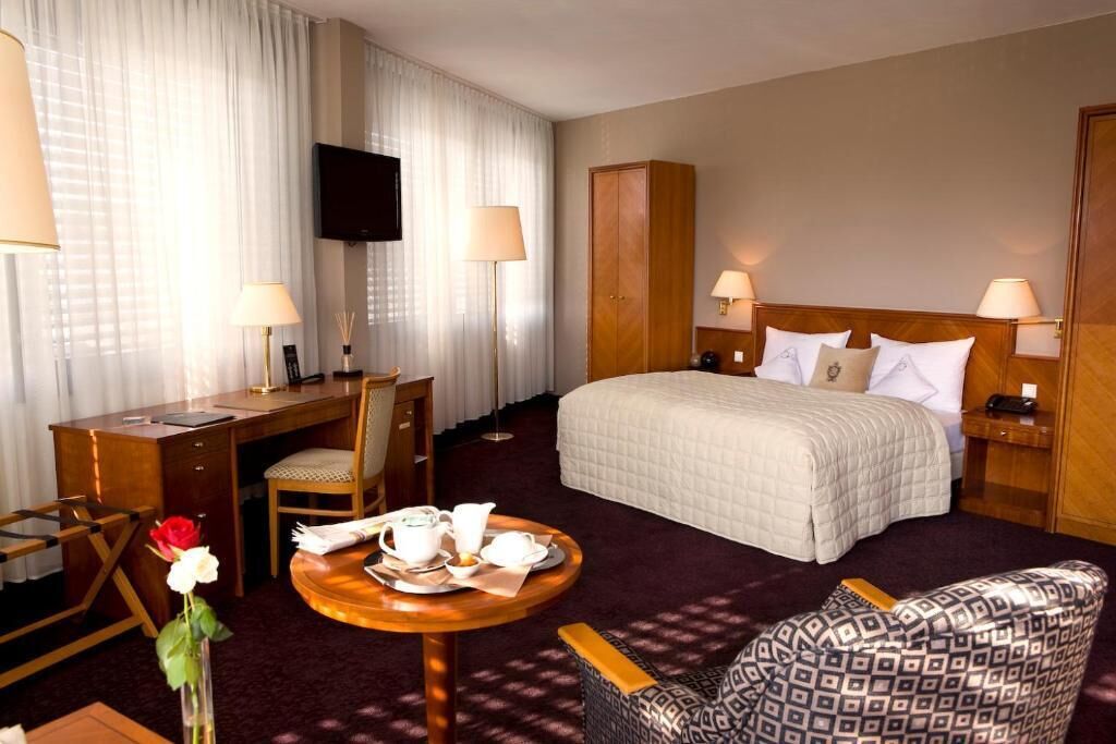 Une chambre d’hôtel avec un lit, un bureau, une chaise, une petite table ornée d’un vase de fleurs et un téléviseur fixé au mur.